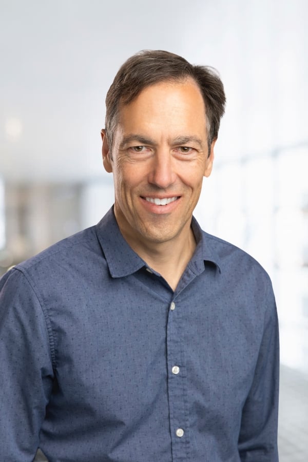 Photo of the top half of a white man with dark hair wearing a blue button-down shirt.
