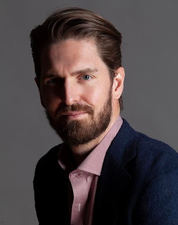 Lee Bradshaw, a light-skinned man with brown hair and a beard, wearing a blue blazer over a pinkish-red collared shirt.