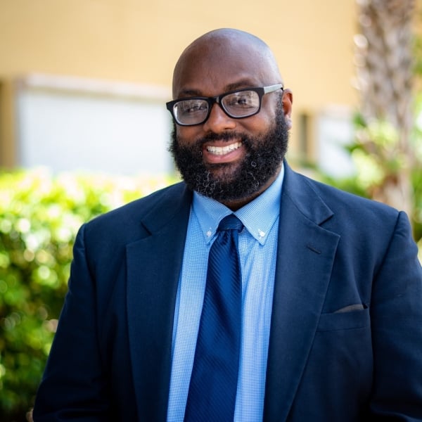 Ronnie Mack, a dark-skinned man with a beard but no hair on his head, wearing glasses and a blue suit.
