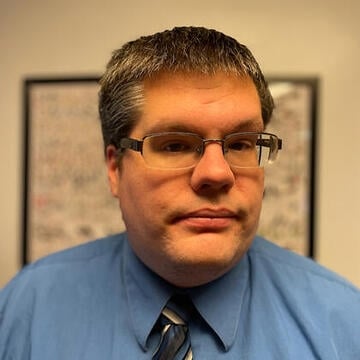 A light-skinned man with short hair and glasses, wearing a blue collared shirt and tie.