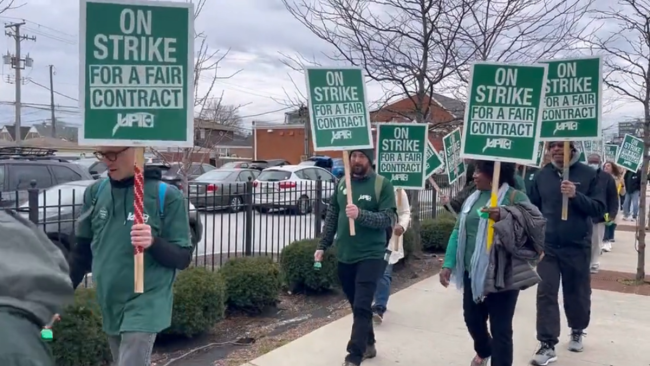 Demonstrators walk on a sidewalk next to a parking lot holding green signs that say "On Strike For a Fair Contract"