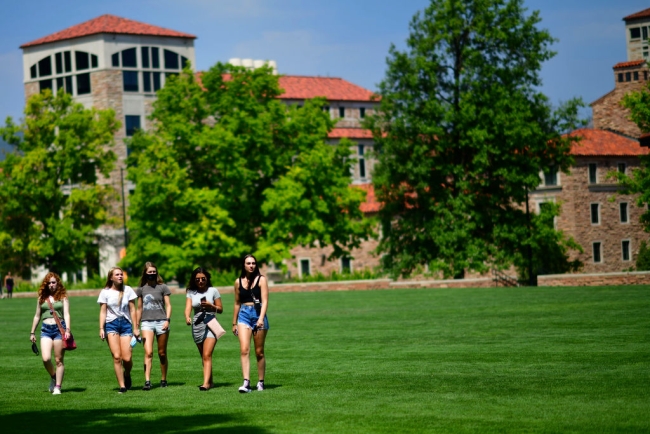 A group of female students walk across a bright green field in front of a brick campus building