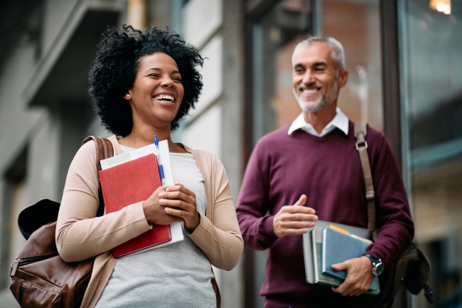 A woman and man, older than traditional college students, laugh while carrying books and wearing backpacks.