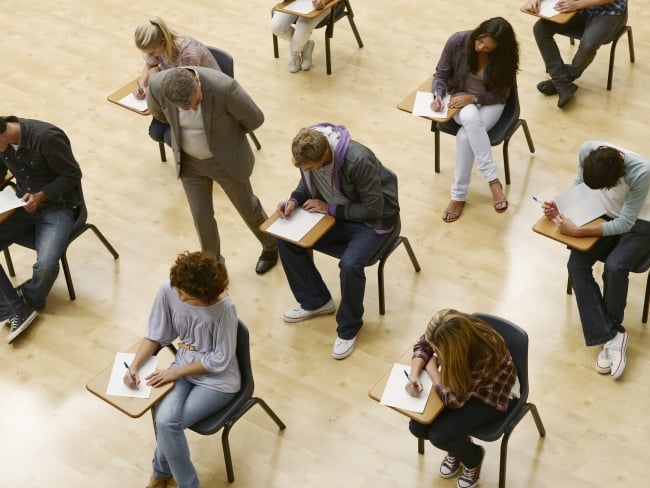 Students sit in a classroom taking a test as a teacher walks by.