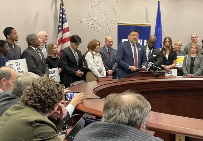 A man in a suit addresses lawmakers backed by supporters