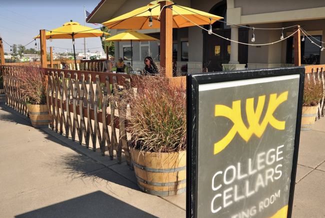 Two people sit in the background of a winery patio under a yellow umbrella. Sign in front of frame says College Cellars.