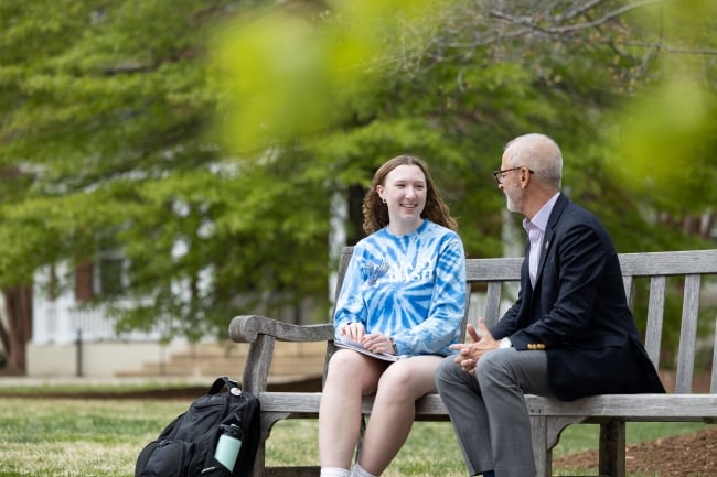 President Paino sits on a campus bench, talking to a student whose backpack is on the ground.