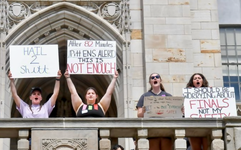 Four Pitt students stand on a balcony holding placards protesting the administration's slow response in notifying them of a reported active shooter threat.
