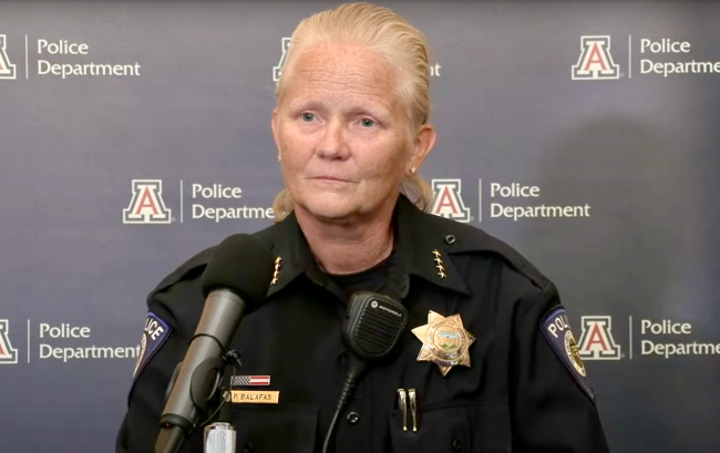 Paula Balafas, the now former chief of the University of Arizona Police Department, stands at a mic with the university logo on a wall behind her. She is a light-skinned middle-aged woman with blond hair.