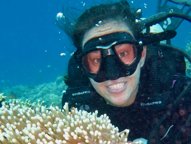 In an underwater photo, a woman in a scuba mask smiles while next to a coral reef