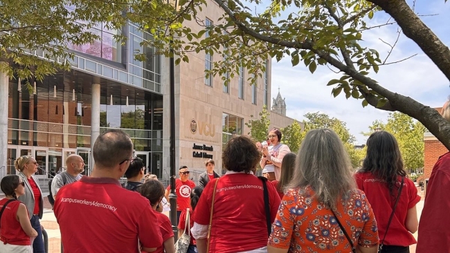 People, mostly in red shirts, some with #campusworkers4democracy on the back, listen to a speaker with a megaphone outside a Virginia Commonwealth University building.