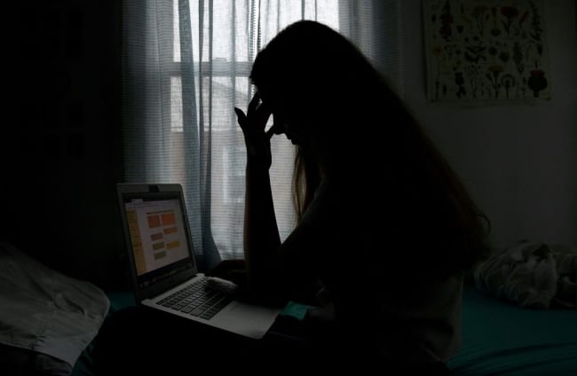 Young woman sits in a dark room in silhouette looking at her open laptop.