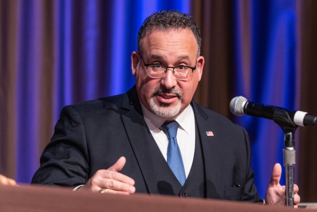 Miguel Cardona, a middle-aged Hispanic man with a goatee and glasses, stands at a podium in a black suit and blue tie.