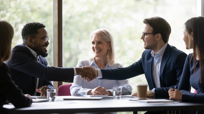 Four diverse people sit around a table, while one man stretches out a hand to welcome another