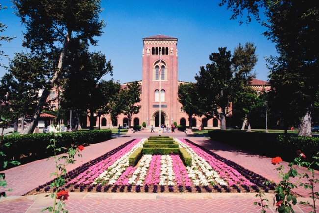 A University of Southern California campus photo, depicting a long walkway toward the entrance of a USC building. A garden with pink and white flowers and well-manicured shrubbery bisects the brick walkway.