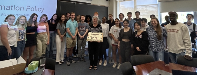 A group of college students surrounding their instructor, in front of a screen with the name of the course on it.