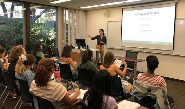 A Scripps College alumna teaches a small class, with a projector screen stating that the lesson is about the three types of budgeting.