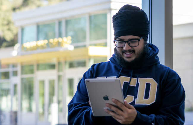 A male student wearing a UC Davis sweatshirt stands by a window in the daytime, looking at his e-reader and presumably reading digitally delivered course materials.
