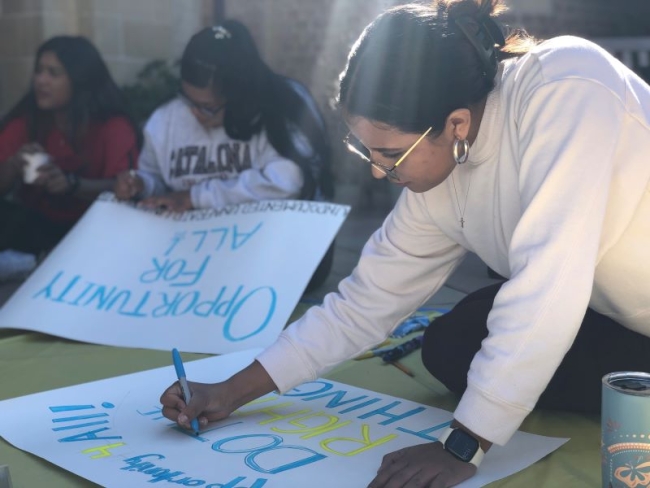 A student writes on a protest sign. Behind her, a student writes on a sign that says "opportunity for all" in blue letters.