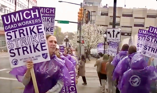 People, most wearing purple ponchos, hold purple and white signs on a sidewalk. The signs say "UMich Grad Workers Strike Strike Strike" and the union's name.