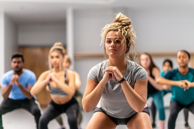 Photo of young people in a fitness class, squatting. Focus is on a young woman in front with her hair in a bun.