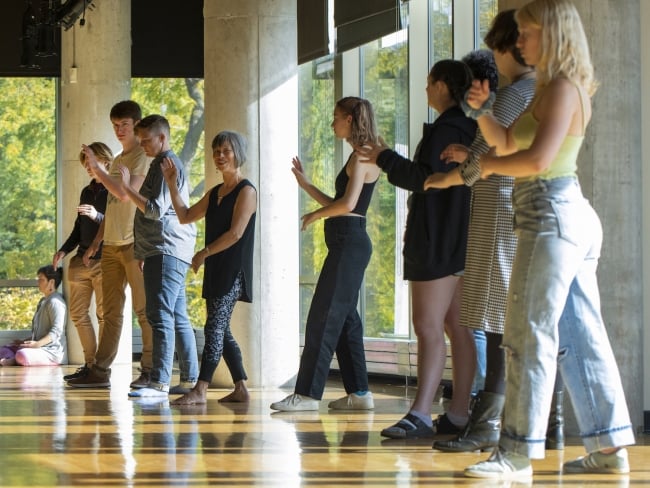 A group of nine people stand in a line practicing tai chi in a well-lit studio