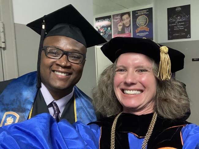 A Black male student in a graduation gown and mortarboard stands next to a light-skinned woman with gray hair who is also wearing academic regalia.