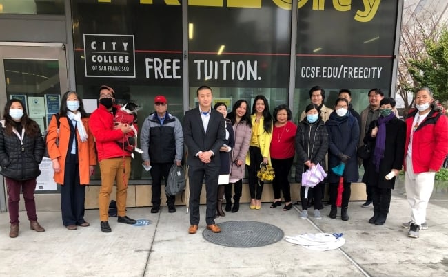 City College of San Francisco board president Alan Wong stands in a row with a group of representatives of organizations supporting Cantonese programming in front of a college building on the Chinatown campus. Many of the people are Asian American, and some are wearing blue paper masks.