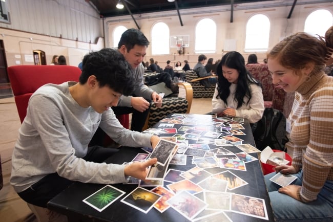 Four students sit around a table looking at dozens of printed-out photographs and images.