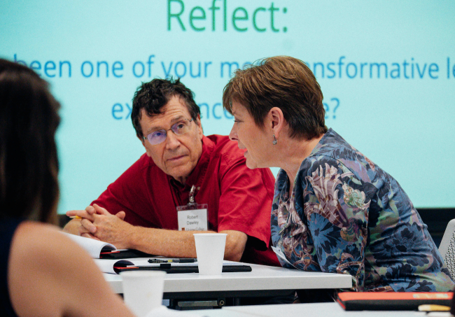 Attendees of a conference, two middle-aged light-skinned people, talk at a table. 