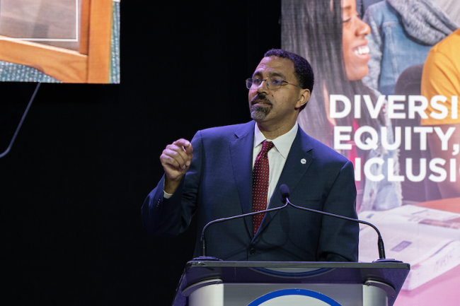 SUNY chancellor John B. King, a dark-skinned man wearing a suit and glasses, is pictured here standing at a podium on a stage in front of colorful images.