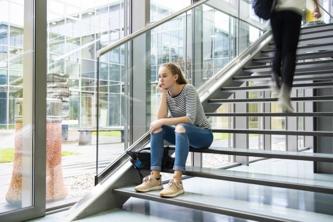 A female college student sits alone in a stairwell, her chin in her hand, as another student walks up the stairs away from her in a blur.