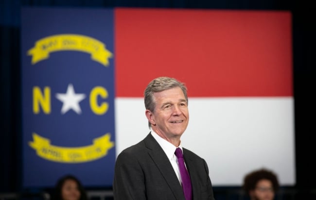 A light-skinned man in a suit and tie with gray hair stands in front of the North Carolina state flag