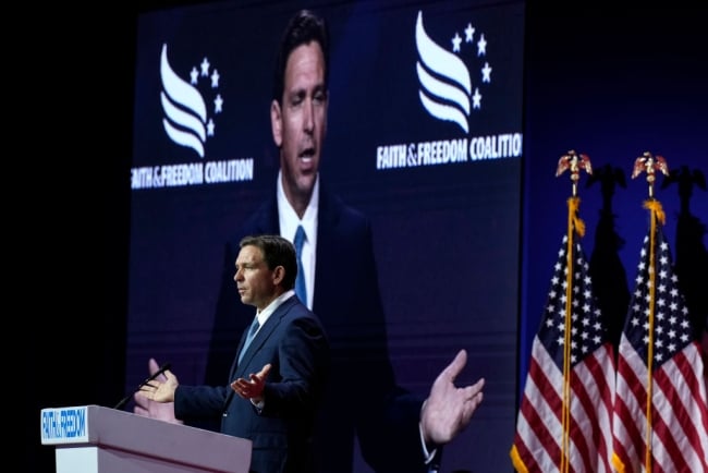 Florida governor Ron DeSantis stands at a podium in front of two American flags. His image is displayed on a big screen behind him.