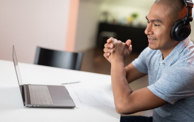 Man with headphones sits a desk with his laptop open as part of a teletherapy session. 