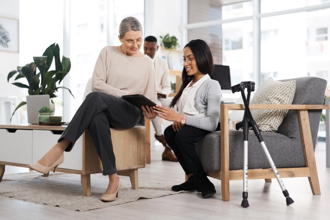 A business woman sits on a chair, with a mobility aid beside her, and speaks with another woman, also sitting.