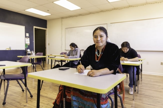 A female Navajo high school student sits at a desk in a classroom, smiling at the camera, her pencil poised over an exam.