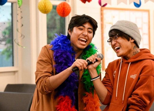 Two students sing karaoke surrounded by rainbow decorations, one of them wearing a rainbow boa