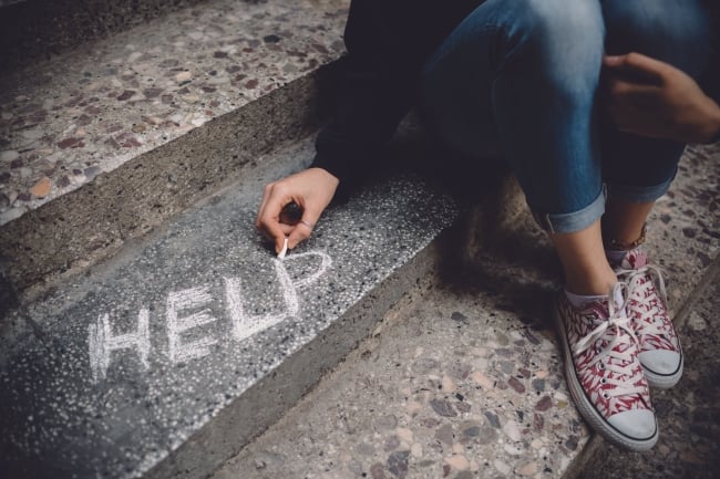 Cropped photo of a student writing "help" in chalk on a set of steps while sitting. Face is not visible.