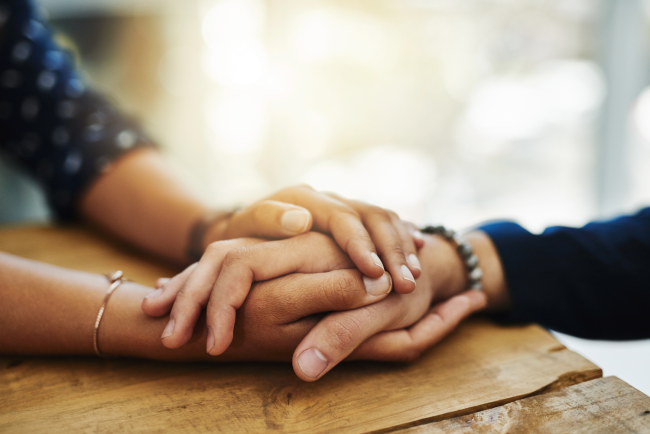 A pair of hands with darker skin resting on a table hold a hand with lighter skin in a comforting manner