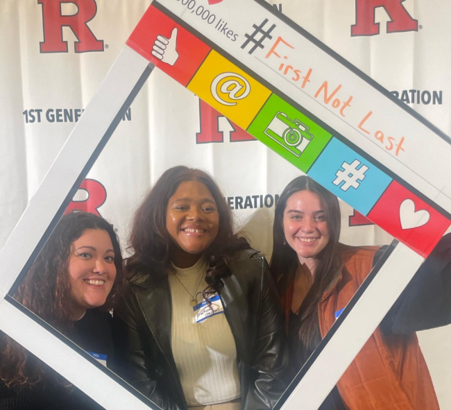 Three first-generation students smile at a Rutgers University first-gen event with a photo-booth frame