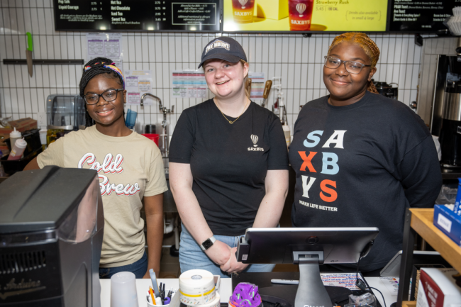 Three students wearing Saxbys shirts smile behind the register in the coffee shop