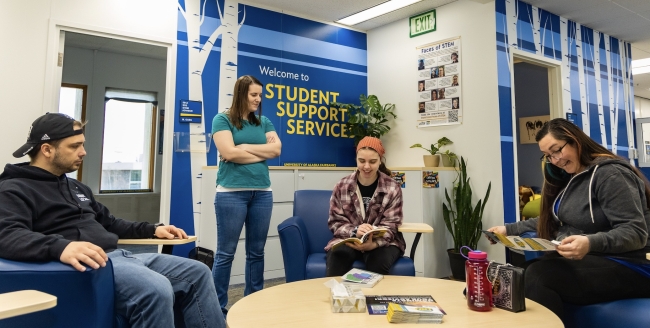 Three seated people in casual clothes speak with a woman standing in a warm-looking campus office space. A large wall sign says "Welcome to Student Support Services."