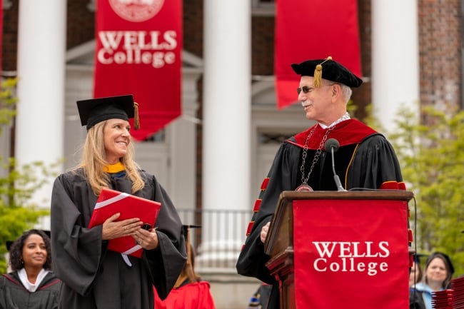 Wells College president Jonathan Gibralter, a light-skinned man with gray hair and a mustache, wearing academic regalia and sunglasses, on graduation day in front of red Wells College signage.