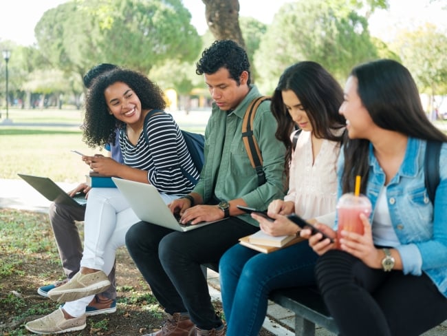 Five students sit outside on a college campus on their laptops and phones.