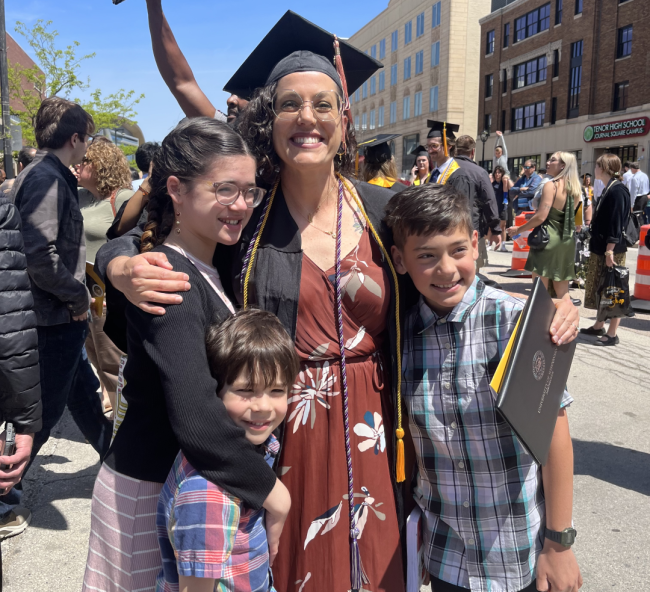 Student Rachel Goochey smiles in her graduation regalia, hugged by her three children.