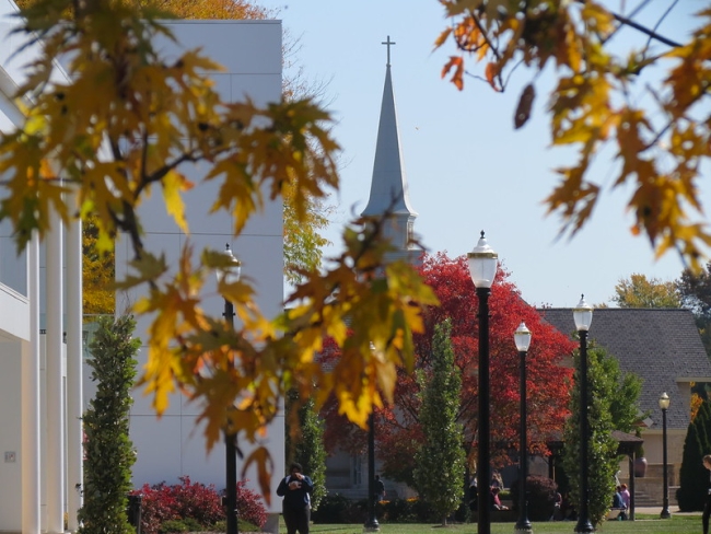 Students walk on Walsh University's campus in the fall with a steeple topped with a cross in the background.