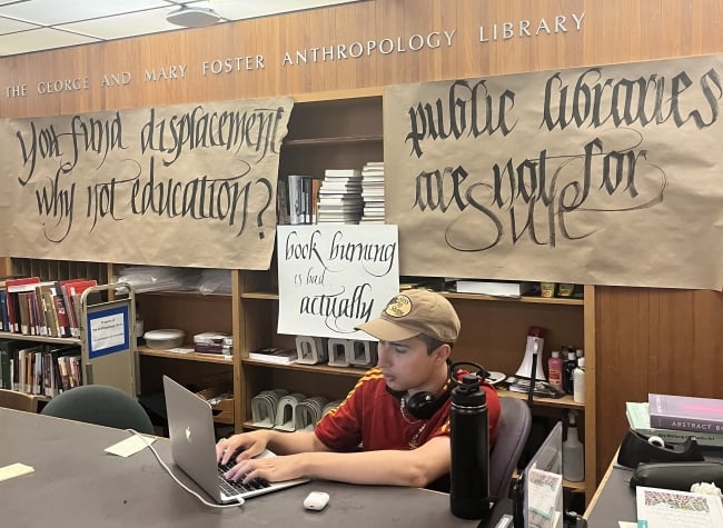A student in a ball cap sits at a laptop with the words "The George and Mary Foster Anthropology Library" on official lettering on the wall behind him, and, on banners below those letters, “You fund displacement why not education?” and “public libraries are not for sale."