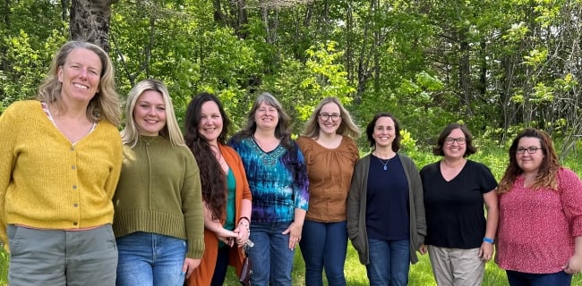 College and career success coordinators from Maine community colleges stand together smiling at a summer retreat with trees in the background.