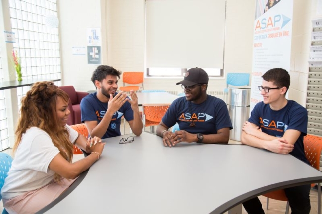 Four students sit around a table talking. Three of them are wearing navy blue shirts that read "ASAP" in blue and orange letters. 